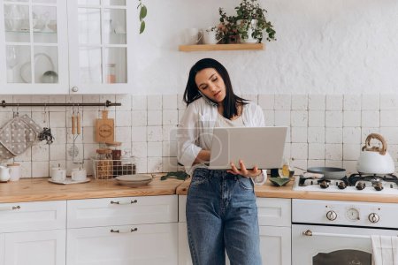 Junge Frau beim Multitasking in der gemütlichen Küche, ferngesteuert am Laptop und telefonierend, umgeben von stilvollem Küchendekor. Konzept der Vereinbarkeit von Privatleben und Arbeit und Fernarbeitsplätze.