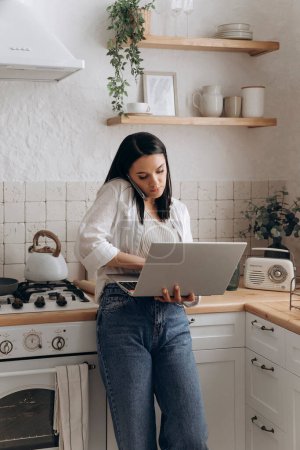Confident young woman freelancer successfully balancing remote work and home chores, talk on the phone call and work on laptop in a well-organized, stylish kitchen. Concept of lifestyle management.