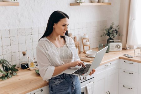 Portrait of young woman freelancer or student working on a laptop in her kitchen, taking breaks between household chores or cooking. Perfect for content about balancing education, work, and home life.