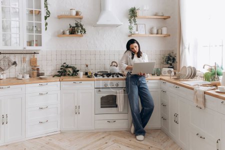 Photo for Confident young woman freelancer successfully balancing remote work and home chores, talk on the phone call and work on laptop in a well-organized, stylish kitchen. Concept of lifestyle management. - Royalty Free Image