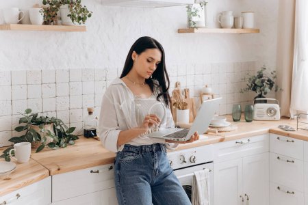 Porträt einer selbstbewussten Freiberuflerin oder Studentin, die in ihrer Küche am Laptop arbeitet, Pausen zwischen Hausarbeit oder Kochen einlegt. Konzept zur Vereinbarkeit von Ausbildung, Arbeit und Privatleben.