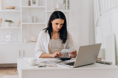 Young woman confidently handling bills and documents at desk with laptop and calculator. Writing notes while checking papers. Managing utility bills, taxes, invoices in well organized white office.