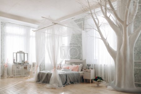 Light-filled bedroom with white canopy bed, pink accent pillows, ornate vanity, artistic white tree, bouquet of flowers, sheer curtains, serene and inviting atmosphere, perfect for relaxation.
