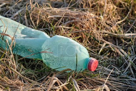 Photo for Crumpled plastic bottle covered with ice and snow in frozen dry grass. Green with red bottle cap. Plastic waste left in nature. Close-up photo. - Royalty Free Image