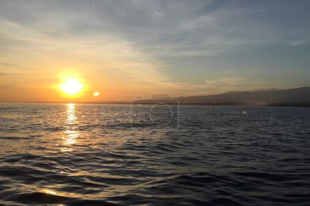 Ein atemberaubender Blick auf den Sonnenuntergang über dem ruhigen Ozean in Bali, Indonesien, mit Bergsilhouetten und goldenen Lichtreflexen.