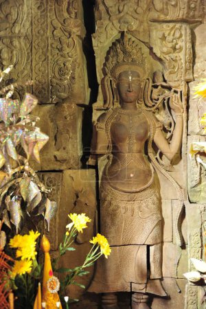 Eine wunderschön detailliert geschnitzte Steinstatue in Angkor Wat, inmitten lebendig gelber Blumen, die die komplizierte Handwerkskunst und spirituelle Aura dieses kambodschanischen Kulturerbes einfängt.