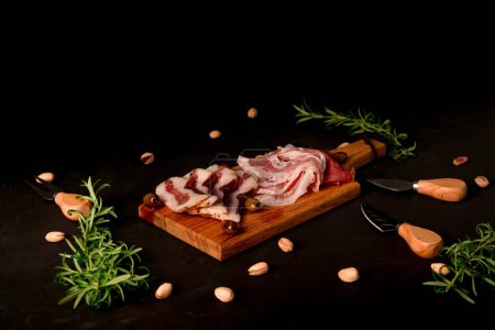 Famous Italian delicacies on wooden board and black background. Guanciale and Speck. delicious Italian hams, olives and pistachios.