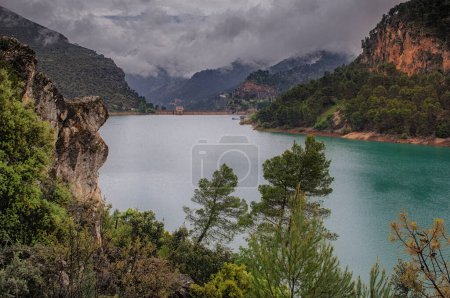 Foto de Embalse de agua rodeado de montañas y cielo nublado en España con pinos en la imagen - Imagen libre de derechos