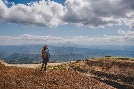 Foto de Mujer viajera y excursionista mirando el paisaje de montaña de verano desde el pico más alto. Vista posterior de la joven con mochila descansando en la parte superior y disfrutando de la escena natural en la cordillera de Kopaonik. - Imagen libre de derechos