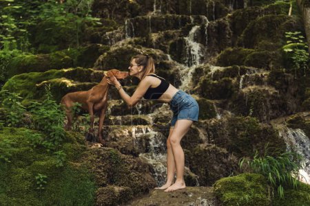 Photo for Young woman with her dog by mountain river waterfall. Female petting Hungarian Vizsla on rock in front of mountain waterfall. Barefoot girl in playing with pet in nature, dog-friendly travel concept. - Royalty Free Image