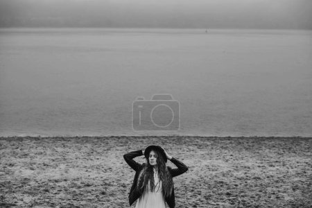 Téléchargez les photos : Young pensive woman in fedora hat standing alone by river bank with eyes closed. Black and white lonely female lost in thoughts on a cold gray day. Monochrome photo with fall melancholy mood concept. - en image libre de droit