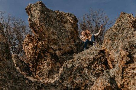 Photo for Mother and child sitting together on forest rock, view from below. Happy family spending weekend in nature. Loving mom showing her excited son something far away sitting on cliff edge on sunny day. - Royalty Free Image