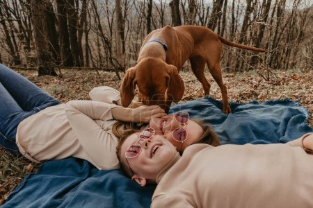 Photo for Mom and son having fun lying on plaid in nature, woman laughing while Vizsla dog licking her face. Mother and child with dimples on cheeks in pink sunglasses. Family together with pet at forest park. - Royalty Free Image