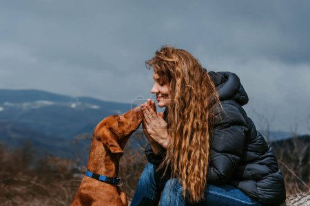 Photo for Young woman traveling with dog. Beautiful female sitting together with Hungarian Vizsla and looking at each other. Happy girl with long curly hair interact with pet at hiking rest with mountain view. - Royalty Free Image