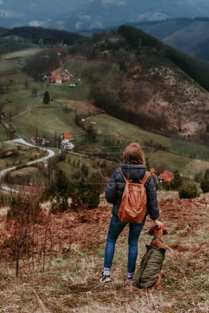 Foto de Mujer joven viajando con perro. Mochilera y Vizsla húngara en chaquetas de pie en la colina y mirando paisaje de montaña escénico. Chica y su mascota senderismo a lo largo de picos de sendero de los Balcanes. - Imagen libre de derechos