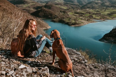 Photo for Travel lifestyle view of young woman and adventure dog. Female backpacker and Hungarian Vizsla sitting on cliff by river above lake. Girl and pet resting and enjoying mountain landscape from rock. - Royalty Free Image