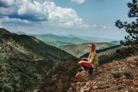 Photo for Woman hiker sitting alone on rock of mountain. Girl traveler overlooks valley with legs crossed, sits on cliff edge and looking away. Female climbed to the top enjoying mountain landscape from peak. - Royalty Free Image
