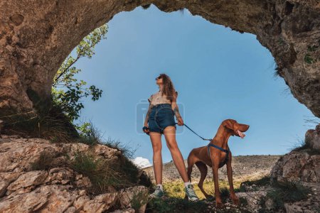 Photo for Young woman with vizsla dog standing in hole in rock. Female travel with pet in nature and climbing to mountain cave. Solo female slow travelling with pointer dog, hiking adventure in wild landscape. - Royalty Free Image