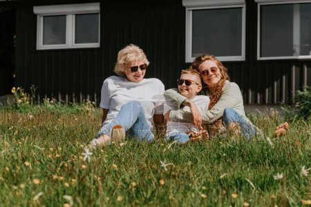 Photo for Grandmother with adult daughter and grandson relaxing outdoors. Happy multi generation family sitting on grass near modern country house, spending time on holidays together. Family vacation concept. - Royalty Free Image