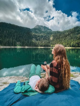 Photo for Mother and child on summer vacation sitting by Black lake in Durmitor national park. Woman pouring tea from thermos while her kid resting on throw blanket, back view. Family enjoying moments together. - Royalty Free Image