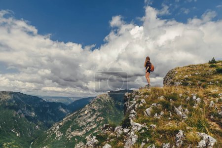 Foto de Mujer mochilera parada sola en la roca de la montaña sobre el cañón del río Tara en Durmitor. Senderista disfrutando de la vista del desfiladero del río Tara desde el borde del acantilado en la cima de la montaña en el Parque Nacional en Montenegro. - Imagen libre de derechos