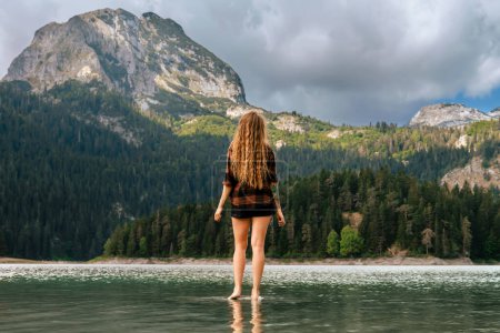 Foto de Mujer parada en el agua y disfrutando de la vista del lago Negro en Durmitor, Montenegro. Mujer en camisa a cuadros se levanta con su espalda y admira el panorama del lago glacial rodeado de bosques silvestres de coníferas. - Imagen libre de derechos