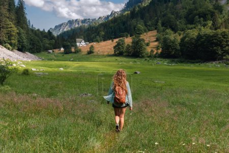 Foto de Mujer con mochila caminando en el prado de montaña de verano en el Parque Nacional Durmitor, Montenegro. Hermosa mujer caminando por el verde valle del lago Susicko, completamente seco durante el verano. - Imagen libre de derechos