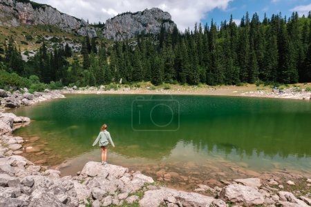 Foto de Mujer de pie sobre una roca en el paisaje de montaña de verano disfrutando de la vista del lago Jablan en Durmitor, Montenegro. Mujer de pie con la espalda y mirando al lago glacial rodeado de bosques de coníferas. - Imagen libre de derechos