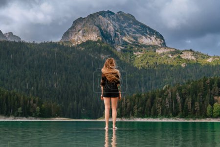 Photo for Female with long flying hair standing in water and enjoying view of Black lake in Durmitor, Montenegro. Woman wearing plaid shirt admires panorama of glacial lake surrounded by wild coniferous forest. - Royalty Free Image