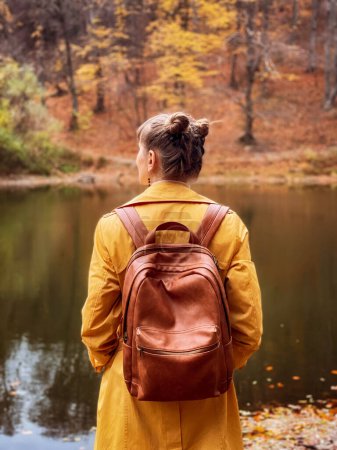 Photo for Back view of woman standing in autumn forest and looking at lake. Young female wearing orange coat standing backwards and watching nature. Girl with brown backpack enjoying fall nature colors in park. - Royalty Free Image