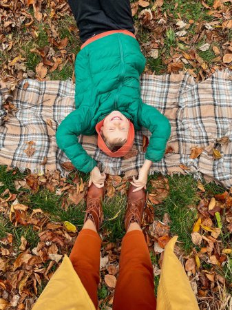 Téléchargez les photos : Vue du dessus de l'enfant heureux riant dans le parc d'automne. Garçon portant une veste verte et un sweat-shirt orange couché sur un pique-nique à carreaux dans les feuilles d'automne et s'amusant. Mère et fils profitant de la journée d'automne sur la nature. - en image libre de droit