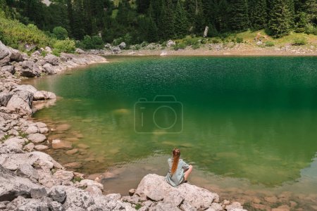 Téléchargez les photos : Femme assise sur un rocher dans un paysage montagneux d'été et bénéficiant d'une vue sur le lac Jablan à Durmitor, Monténégro. Femme assise avec son dos et regardant le lac glaciaire entouré de forêts de conifères. - en image libre de droit