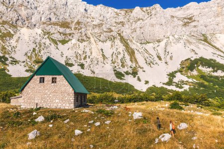 Foto de Paisaje de montaña de verano con cabaña de piedra en el Parque Nacional Durmitor, Alpes Dináricos, Montenegro. Mujer con hijo y perro caminando hacia la cabaña cerca de los lagos glaciares de Skrcko. Gente pequeña en paisaje grande. - Imagen libre de derechos