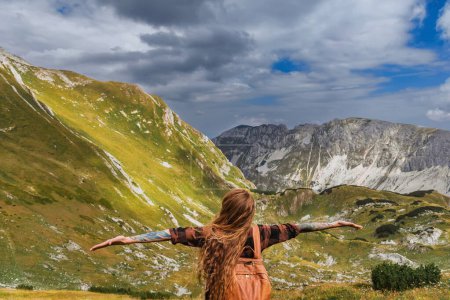 Photo for Woman solo travel stands alone with open arms in mountains. Girl traveler raised hands standing with her back and enjoying mountain landscape. Female exploring natural beauty of Durmitor, Montenegro. - Royalty Free Image
