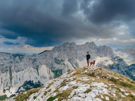 Foto de Hombre activo con perro de pie en la cima de la montaña, descansando después de escalar. Senderista masculino con su Vizsla húngara disfrutando de vistas durante la caminata en el parque nacional de Durmitor. Senderismo para mascotas y concepto de viaje. - Imagen libre de derechos