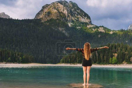 Photo for Young woman enjoying summer vacation by mountain lake, her arms stretched out towards the mountains as symbol of freedom. Woman traveler standing with her back in Black lake, Durmitor, Montenegro. - Royalty Free Image