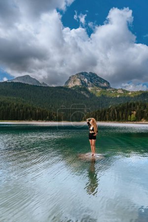 Photo for Young beautiful woman enjoying summer vacation by mountain lake, her arm tucking long curly hair. Woman traveler standing on rock in water of Black lake in National park Durmitor, Montenegro. - Royalty Free Image