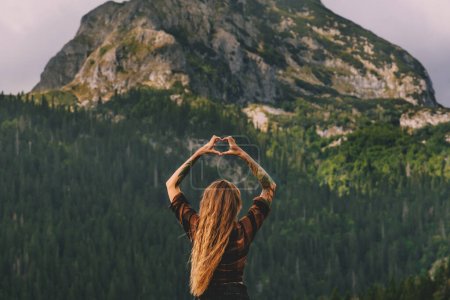 Foto de Mujer joven viajera sosteniendo sus manos en forma de corazón contra el fondo de las altas montañas. Hermosa chica tatuada de pie mirando hacia atrás viajando en el Parque Nacional Durmitor, Montenegro. - Imagen libre de derechos