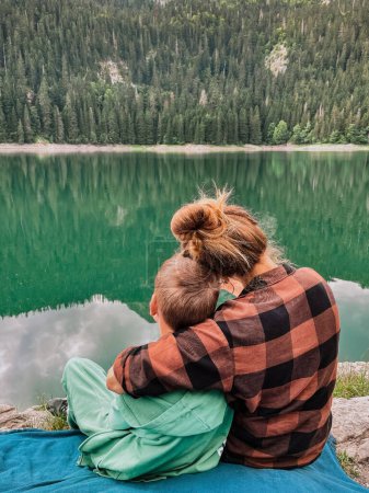 Téléchargez les photos : Mère embrassant son fils assis sur un rocher près du lac noir dans le parc national Durmitor. Maman et son enfant se détendent sur une couverture de pique-nique et regardent l'eau, vue arrière. Heureux famille passer du temps ensemble sur la nature. - en image libre de droit