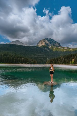 Foto de Mujer de pie en el lago Negro mirando delante de ella disfrutando de la actividad de ocio al aire libre en Durmitor, Montenegro. Mujer de pie sobre roca, admira panorama de lago glacial rodeado de bosque de coníferas. - Imagen libre de derechos