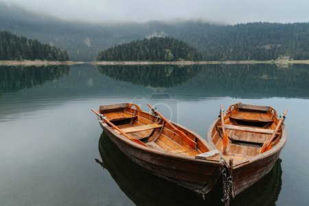 Photo for Two traditional rowing boats on mountain lake covered with mist. Rowboats on calm water of Black lake surrounded by coniferous green forest in foggy morning in Durmitor National park, Montenegro. - Royalty Free Image