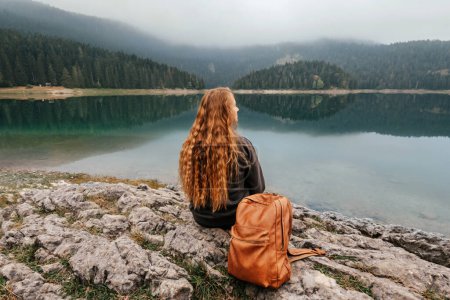 Photo for Woman backpacker sitting alone on rock by mountain lake and enjoying view of Black lake in Durmitor. Female solo traveler meditating enjoying silence and alone time, watching scenery of misty lake. - Royalty Free Image
