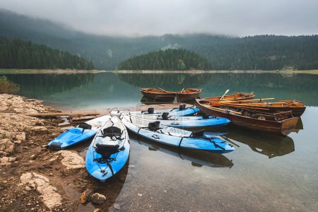 Téléchargez les photos : Bateaux d'aviron et kayaks sur un lac de montagne couvert de brouillard. Bateaux à rames et canots sur les eaux calmes du lac Noir entouré de forêts de conifères dans la matinée brumeuse dans le parc national de Durmitor, Monténégro. - en image libre de droit