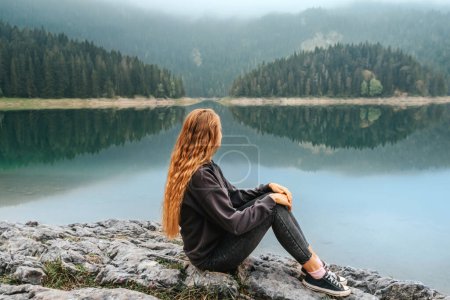 Téléchargez les photos : Jeune belle femme assise seule sur un rocher au bord d'un lac de montagne et bénéficiant d'une vue sur le lac noir à Durmitor. Voyageuse solo relaxante jouissant du silence et du temps seul, regardant des paysages de lac brumeux. - en image libre de droit