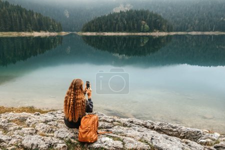 Téléchargez les photos : Une touriste assise seule au bord d'un lac de montagne et prenant des photos avec son téléphone. Voyageuse solo profitant de vues et prend des photos à l'aide d'un téléphone mobile. Scène sur le lac noir brumeux à Durmitor. - en image libre de droit