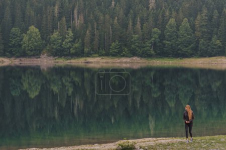 Foto de Mujer caminando a orillas del lago Negro rodeado de bosque de coníferas en el Parque Nacional Durmitor, Montenegro. Caminatas femeninas en la costa visitando el paisaje matutino con el panorama brumoso de Crno Jezero. - Imagen libre de derechos
