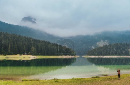 Photo for Woman tourist standing alone by mountain lake and and taking photos with her phone. Female solo traveler enjoying views and takes pictures using mobile phone. Scene on misty Black Lake in Durmitor. - Royalty Free Image