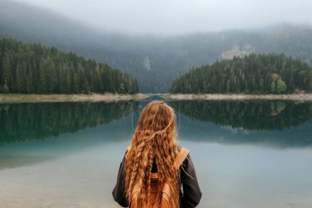 Foto de Mochilero mujer de pie solo en la orilla del lago de montaña y disfrutando de la vista del lago Negro en Durmitor. Viajera solitaria meditando disfrutando del silencio y del tiempo a solas, observando paisajes de lago brumoso. - Imagen libre de derechos
