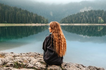 Téléchargez les photos : Jeune femme relaxante, profitant du silence et du panorama touristique du lac noir. Voyageuse solitaire assise seule sur un rocher au bord d'un lac de montagne et méditant, regardant des paysages de lac brumeux à Durmitor. - en image libre de droit