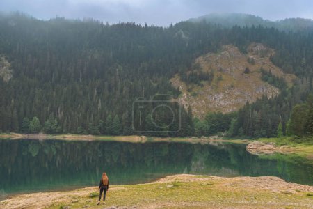 Photo for Woman standing on lake shore of foggy Black lake surrounded with coniferous forest in Durmitor National Park, Montenegro. Young female sightseeing morning landscape with misty panorama of Crno Jezero. - Royalty Free Image